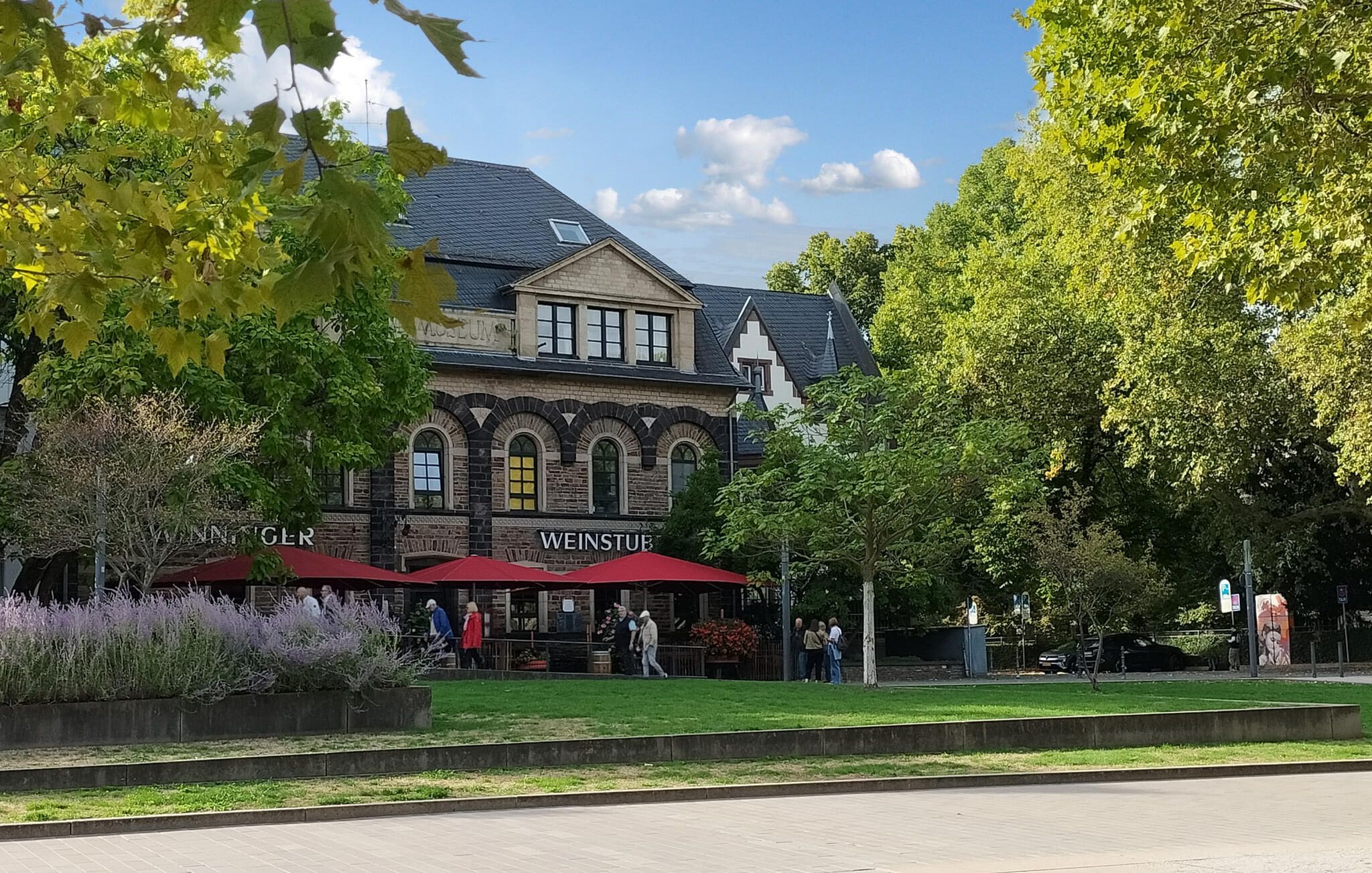 A historic building with a sign reading 'Museum' and 'Wenstube' on the facade, surrounded by trees and a paved area with people.