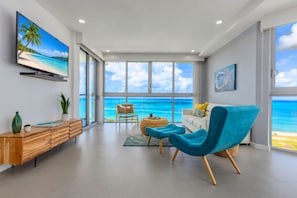 Ein modernes Wohnzimmer mit einem großen Fenster mit Blick auf einen Strand, einem an der Wand befestigten Flachbildfernseher und einem Holzkonsolentisch mit Dekorationen.