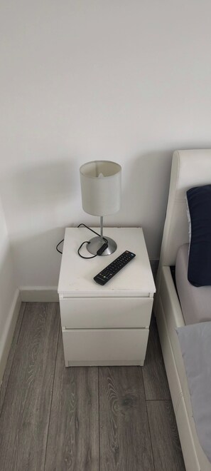 A white bedside table with a lamp, a remote control, and a white nightstand in a room with a wooden floor. - Room-small Double Room in Chelmsford Fre (Chelmsford)