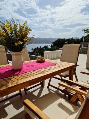 Eine Terrasse im Freien mit einem Holztisch und -stühlen, einer Vase mit gelben Blumen und Blick auf das Meer und die Berge.