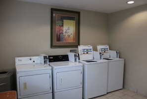 A laundry room with four white washing machines and dryers, a framed picture on the wall, and a trash bin. - Ullah Lodging  (Corpus Christi)