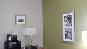 A hotel room with a desk, a lamp, a framed picture, and a coffee maker.