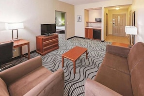Hotellihuone, jossa on sohva, televisio, pieni pöytä, minikeittiö ja kuviollinen matto.