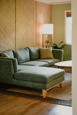 A green sectional sofa with wooden legs, a white table, and a lamp in a room with wooden paneling and a green wall. - The Alani at Kona Shores (Kailua-Kona)