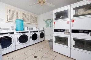 A laundry room with multiple washing machines, a ceiling fan, and a trash bin. - The Alani at Kona Shores (Kailua-Kona)