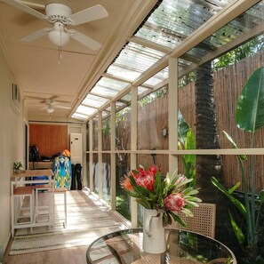 Une pièce baignée de lumière, avec un ventilateur de plafond, une porte vitrée donnant sur l’extérieur et une table sur laquelle est posé un vase rempli de fleurs.