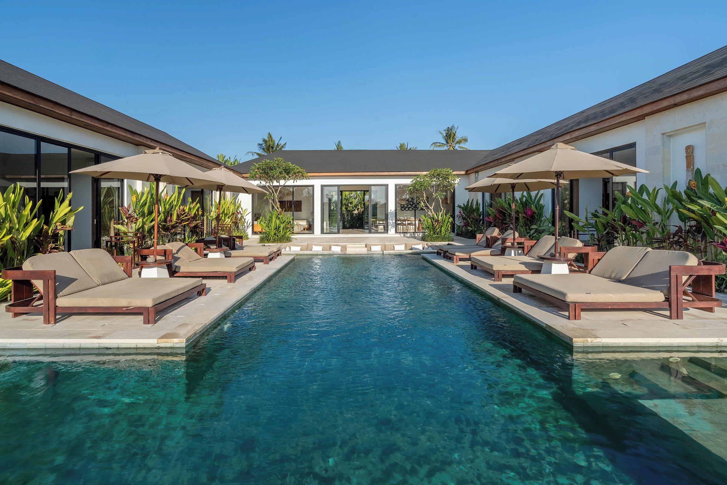 A pool with lounge chairs and umbrellas, surrounded by a building with large windows and tropical plants.