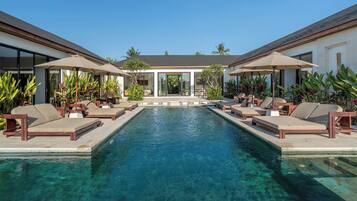A pool with lounge chairs and umbrellas, surrounded by a building with large windows and tropical plants.