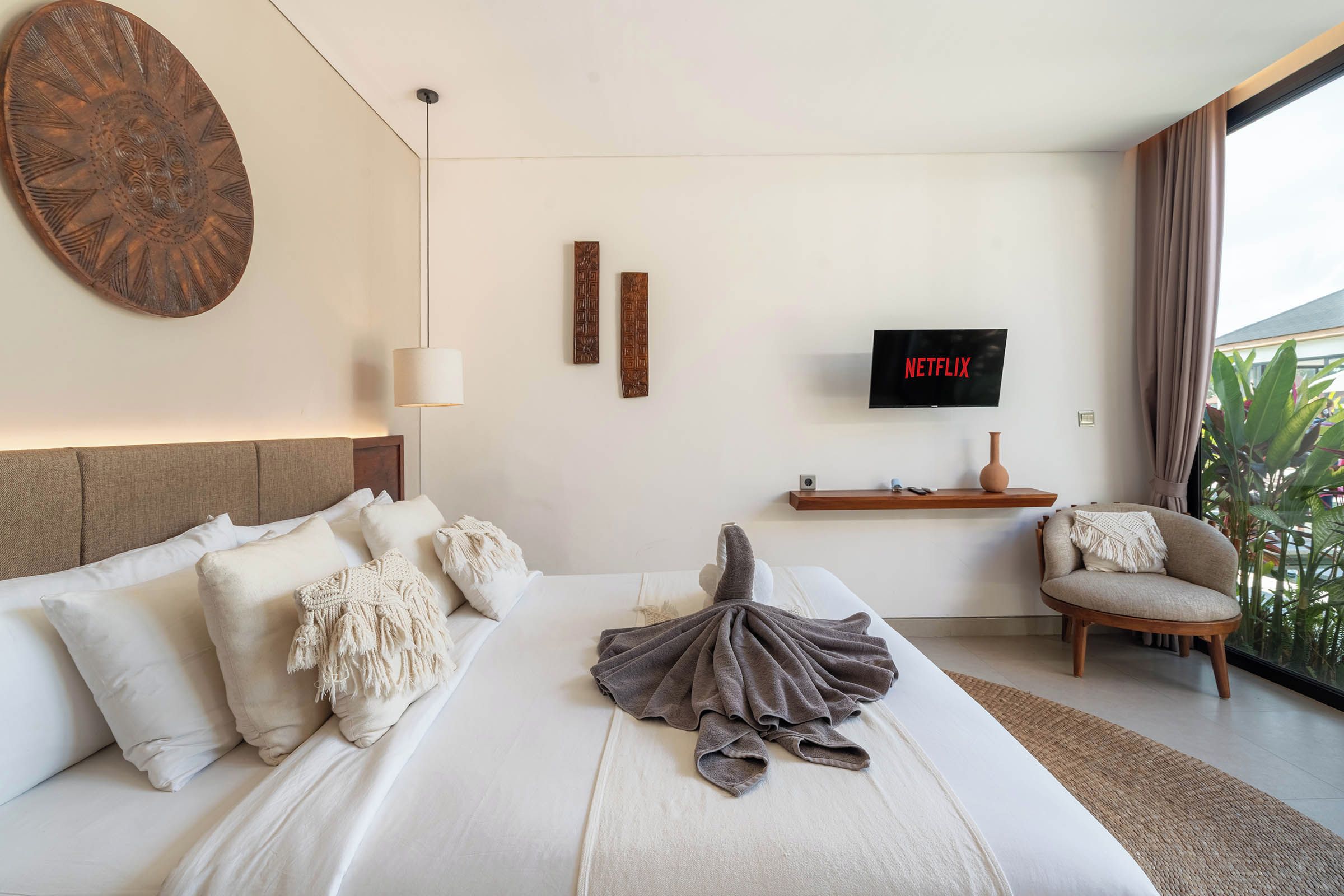 A hotel room with a large bed, a TV mounted on the wall, a small side table, and a view of the outdoors.