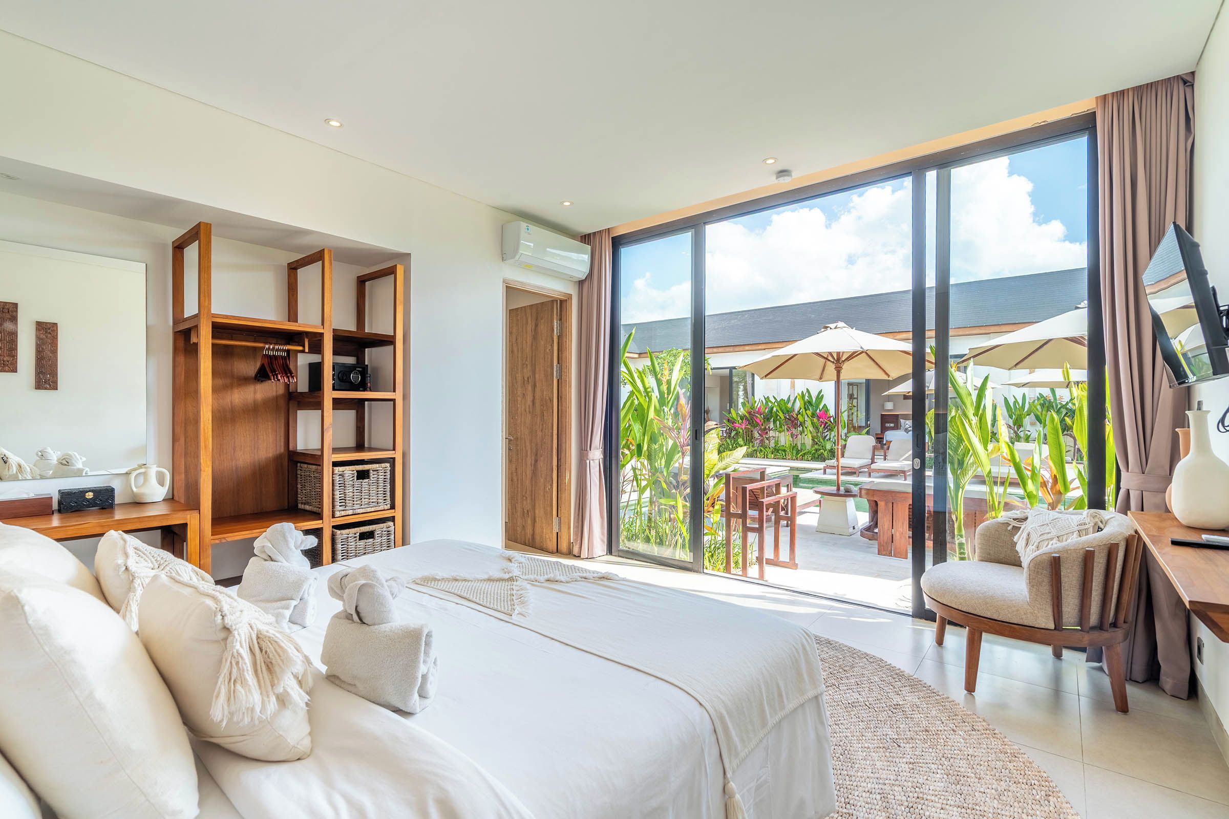 A bedroom with a large bed, a wooden wardrobe, a desk with a chair, and a view of an outdoor patio with umbrellas.