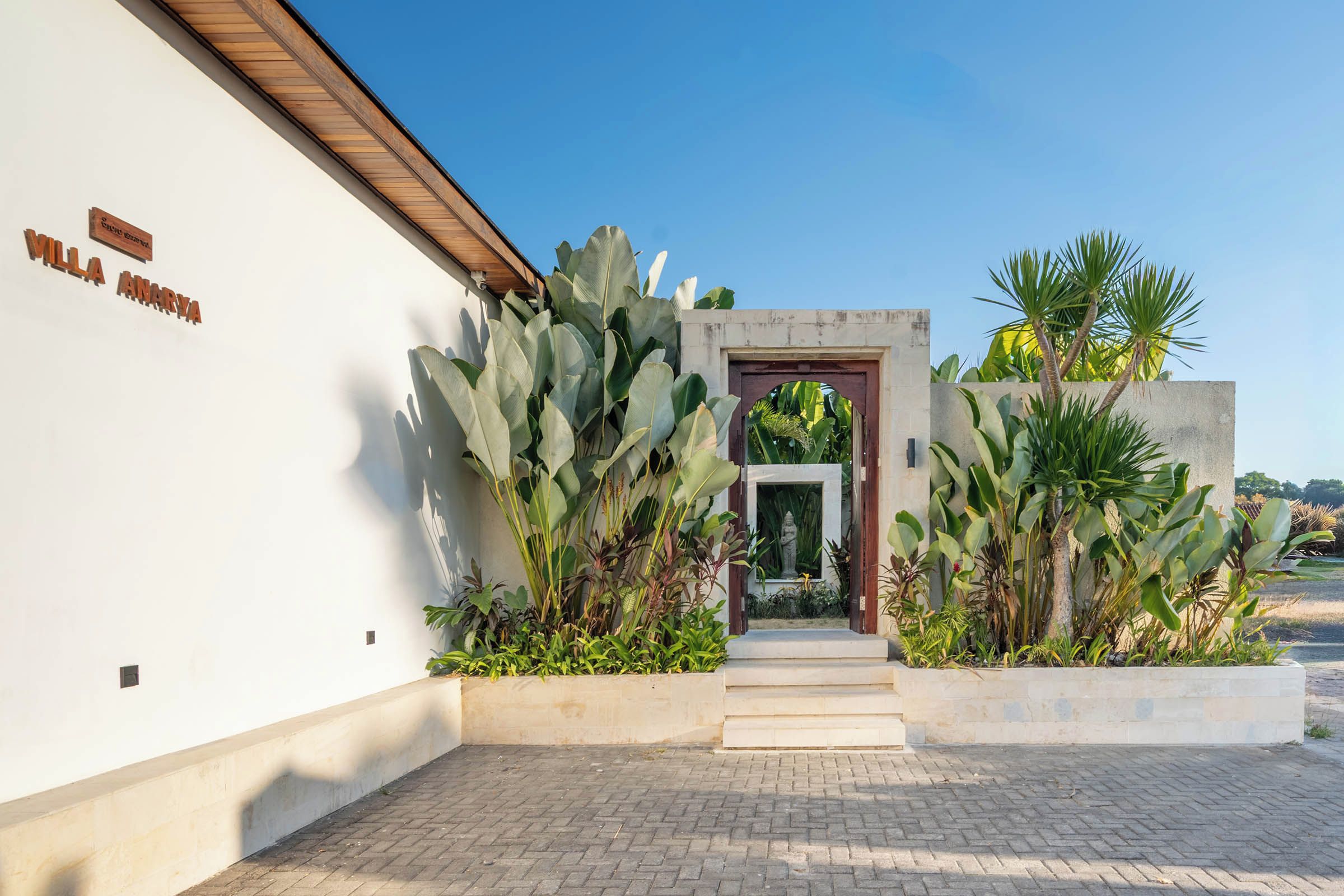 A villa entrance with a wooden door, a nameplate, and a paved pathway leading to a small garden with tropical plants.