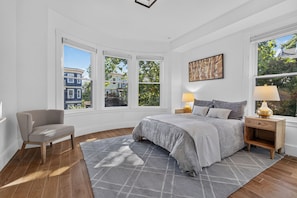 Ein Schlafzimmer mit einem Bett, einem Sessel, einem Nachttisch, einer Lampe und großen Fenstern mit Blick auf Gebäude und Bäume.