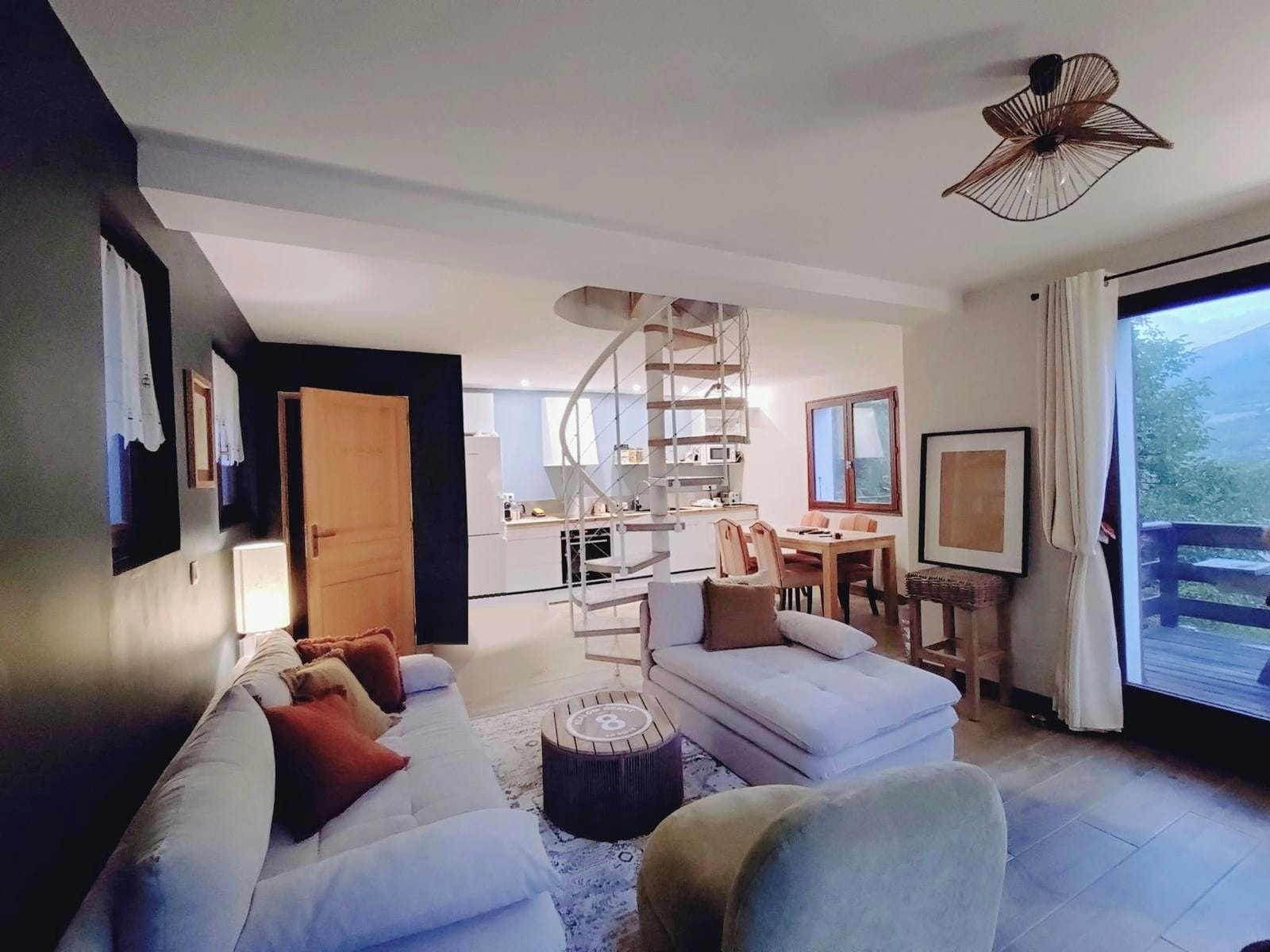 A modern living room with a sofa, ottoman, and a dining area with chairs and a table. A spiral staircase connects the two areas.