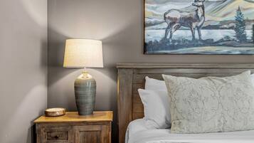 A bedroom with a wooden nightstand, a lamp, a bed with white bedding, and a painting of a deer on the wall.