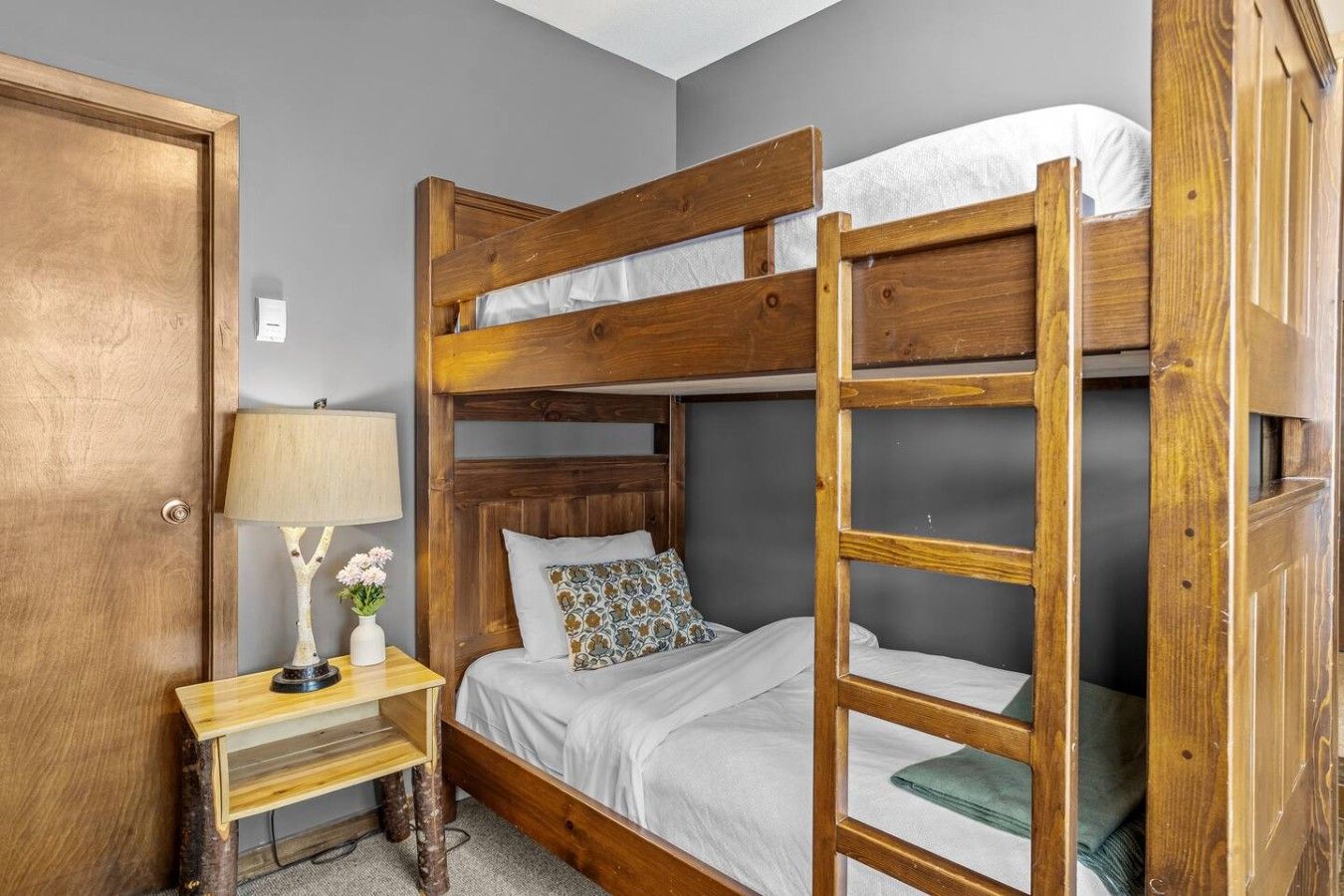 Een stapelbed met houten frame, een nachtkastje met een lamp en een deur.
