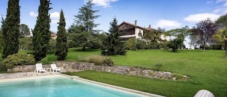 A swimming pool with lounge chairs, surrounded by greenery and a house in the background.