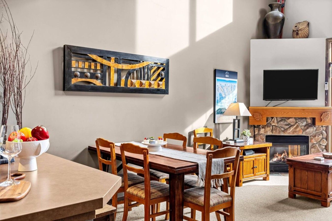 Een eetgedeelte met een tafel, stoelen en een schaal fruit. Er is een open haard met een stenen omlijsting en een aan de muur bevestigde televisie.