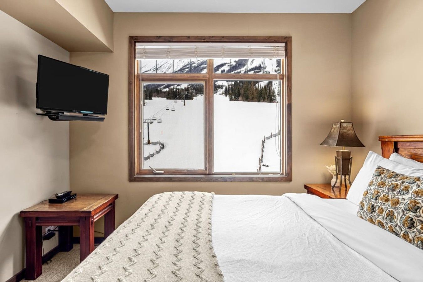 A bedroom with a large window, a bed, a bedside table, a lamp, and a flat-screen TV mounted on the wall.