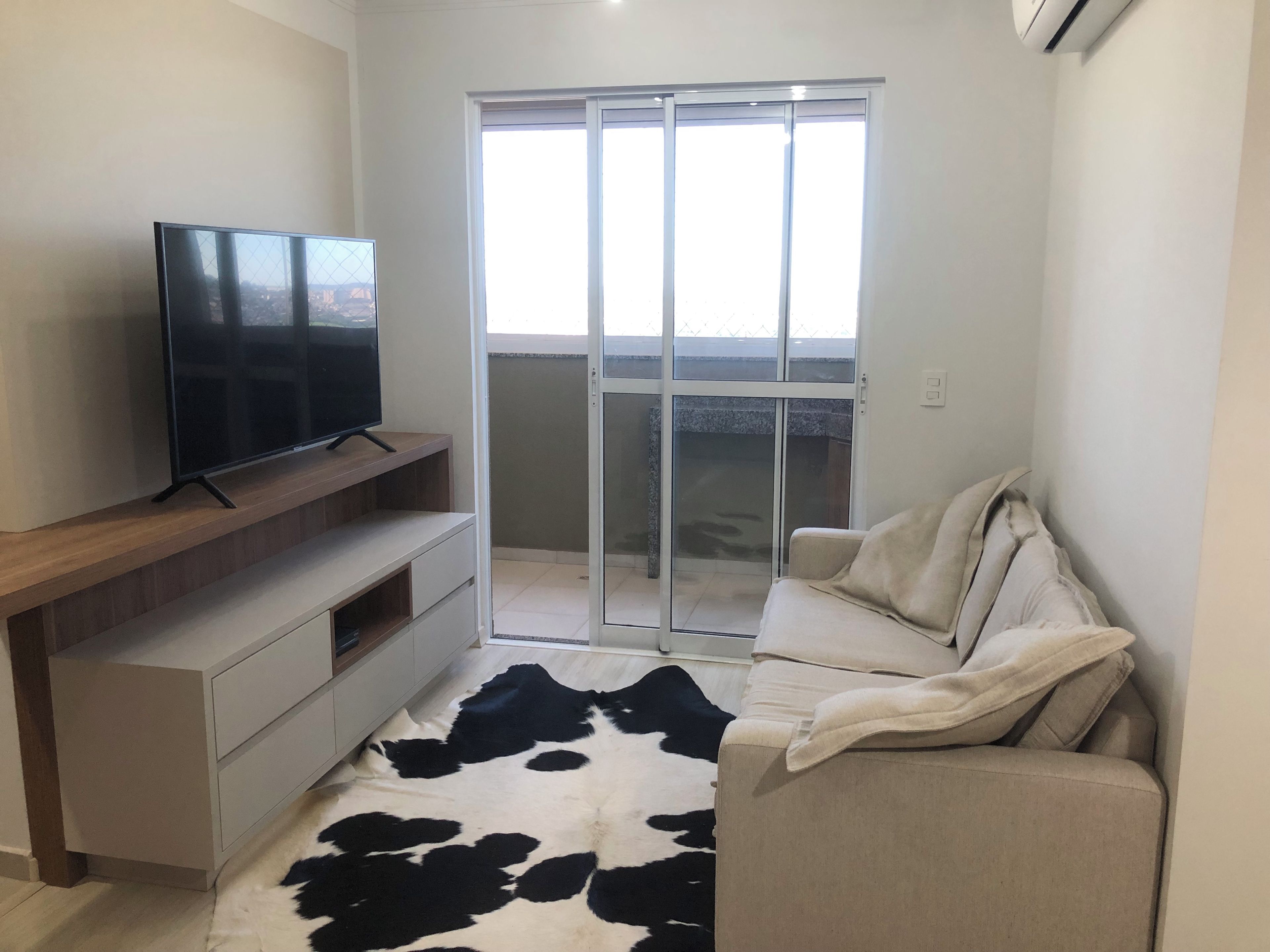 En moderne stue med et fladskærms-tv på et træpanel, en sofa med en ko-skinds måtte, og en skydedør der fører ud til en balkon.