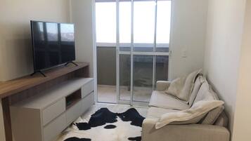 En moderne stue med et fladskærms-tv på et træpanel, en sofa med en ko-skinds måtte, og en skydedør der fører ud til en balkon.