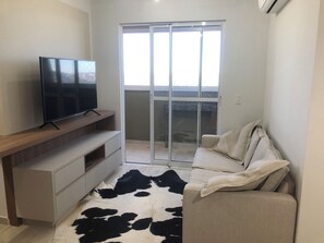 A modern living room with a flat-screen TV on a wooden console, a sofa with a cowhide rug, and a sliding glass door leading to a balcony. - NEW - 100 meters from the JL Shopping Center, garage, 3 elevators, balcony with grill. (Cascavel)