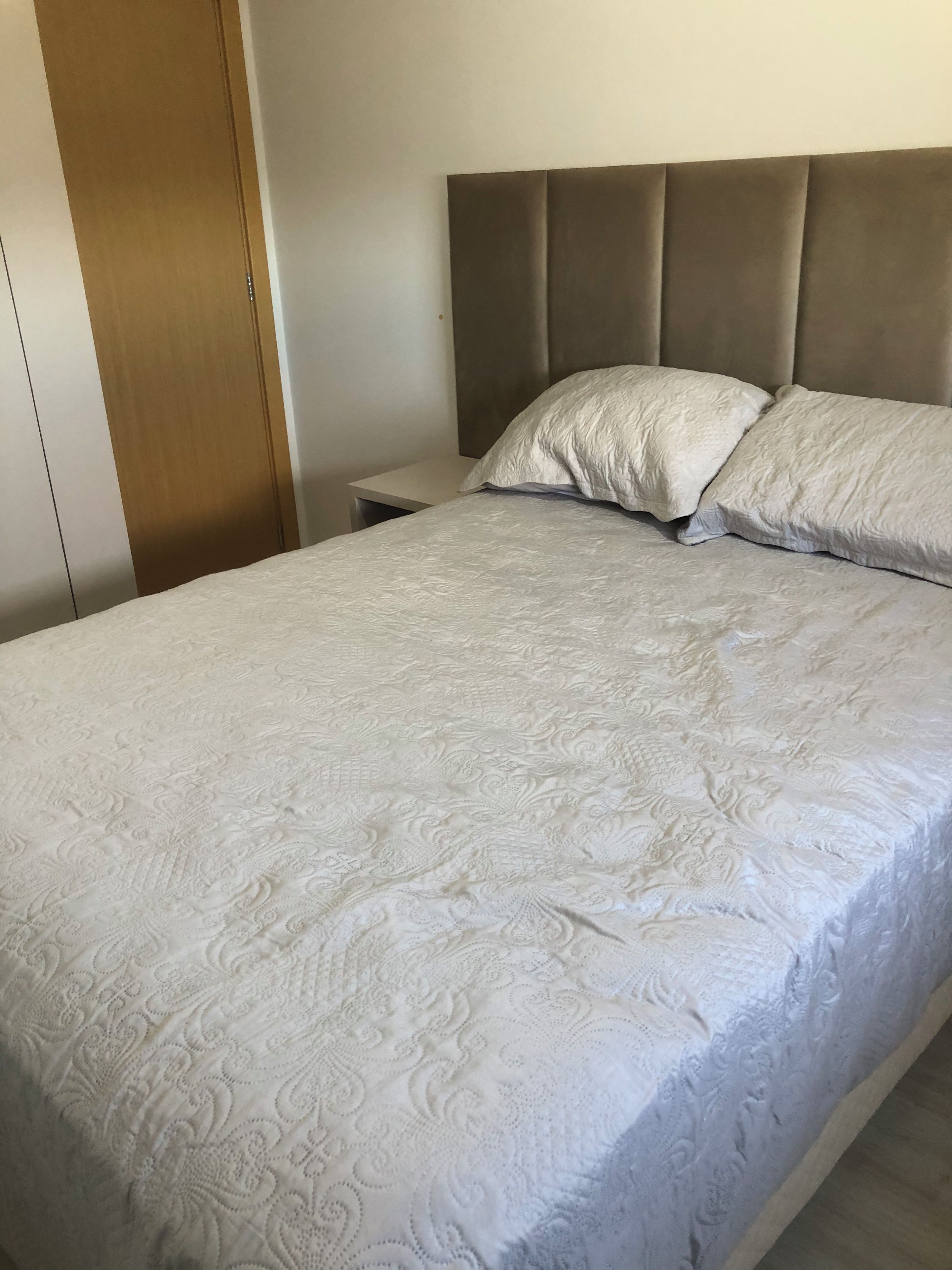 Cama bem arrumada com colcha estampada, guarda-roupa de madeira e mesa de cabeceira.