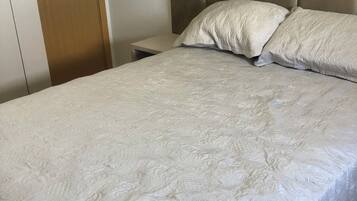 Cama bem arrumada com colcha estampada, guarda-roupa de madeira e mesa de cabeceira.