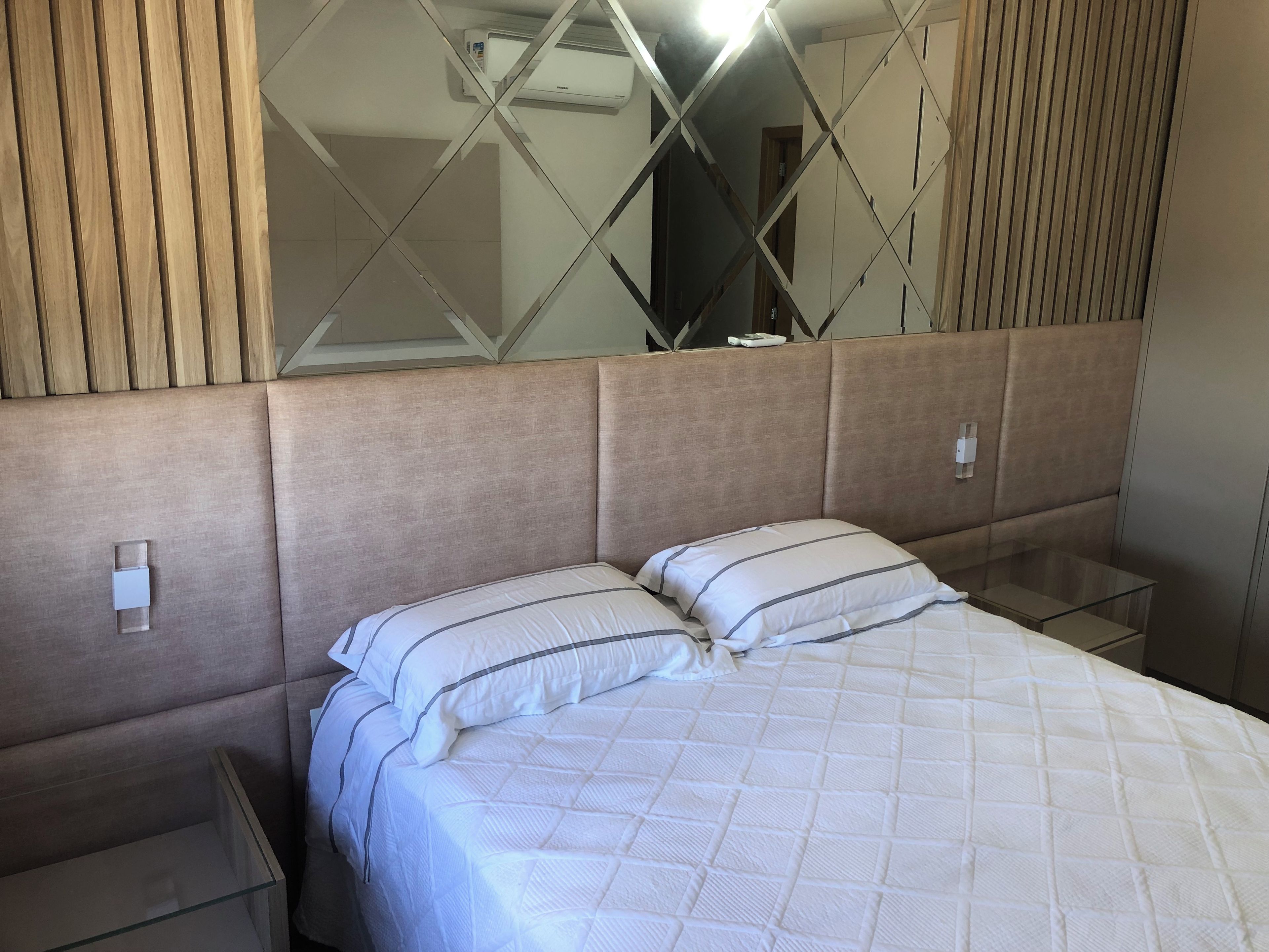 A hotel room with a bed, a nightstand, a mirror, and a wall-mounted air conditioning unit.