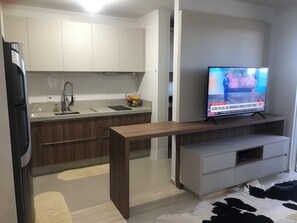A modern kitchen with a built-in dishwasher, a wooden bench, and a TV displaying a news channel. - NEW - 100 meters from the JL Shopping Center, garage, 3 elevators, balcony with grill. (Cascavel)