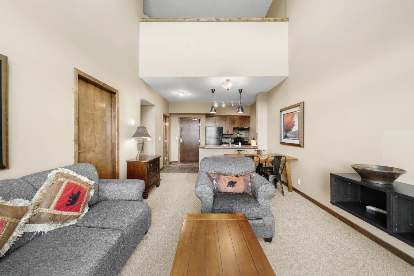 A living room with a sofa, armchair, and a coffee table. There is a kitchen in the background with a dining table and chairs.