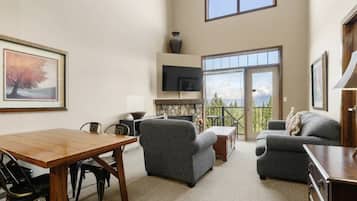 A spacious living area with a dining table, two sofas, a fireplace, and a large window offering a view of the outdoors.