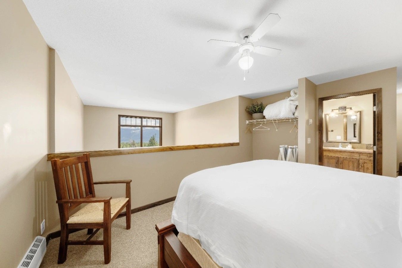 A bedroom with a bed, a chair, a ceiling fan, a window, and a bathroom visible through an open door.