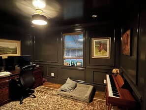 Ein Zimmer mit Klavier, Schreibtisch mit Computer, Stuhl, Sofa, Fenster mit Jalousien und einem Gemälde an der Wand.