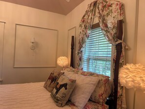 Une chambre à coucher avec un lit, des rideaux à motifs floraux, une fenêtre avec des stores et une lampe fixée au mur.