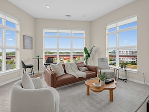 A spacious living room with a brown leather sofa, a white armchair, and a wooden coffee table. Large windows offer a view of a bridge and greenery. - Brand-new Luxury Furnished Penthouse in Noda 30 Night Mid-term Rental (Charlotte)