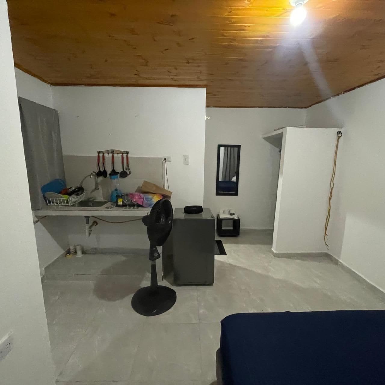 Habitación pequeña con cama, escritorio y silla, un refrigerador compacto y un ventilador.