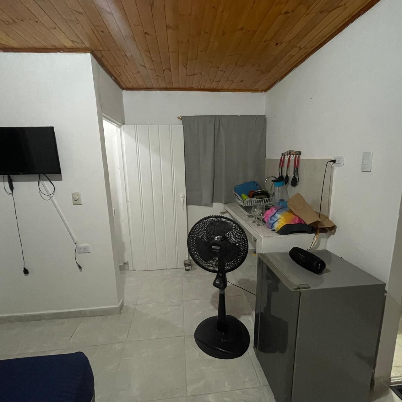 A room with a television mounted on the wall, a standing fan, a small refrigerator, and a window with a curtain.
