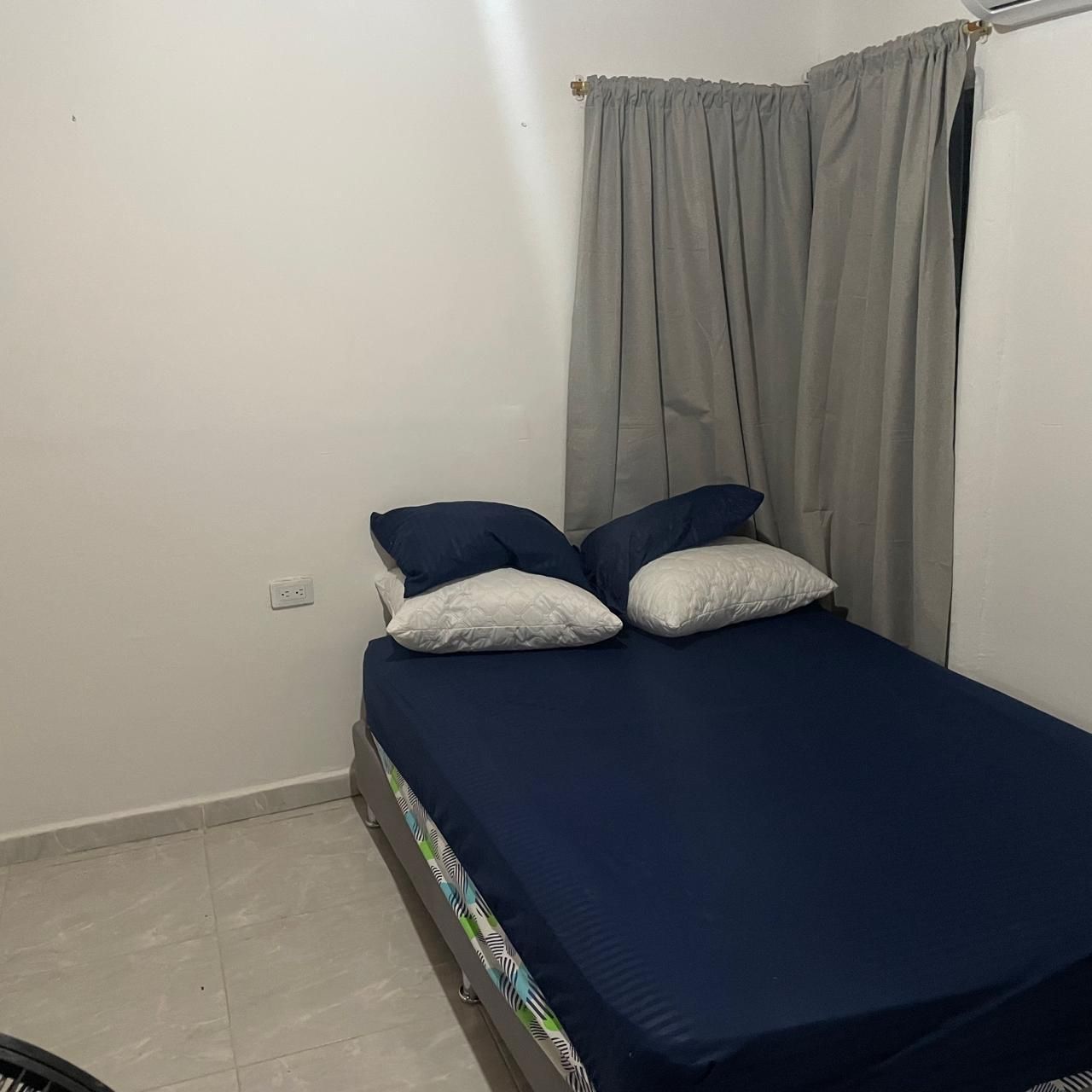A single bed with a blue mattress and two pillows, a grey curtain, and a white wall with a power outlet.