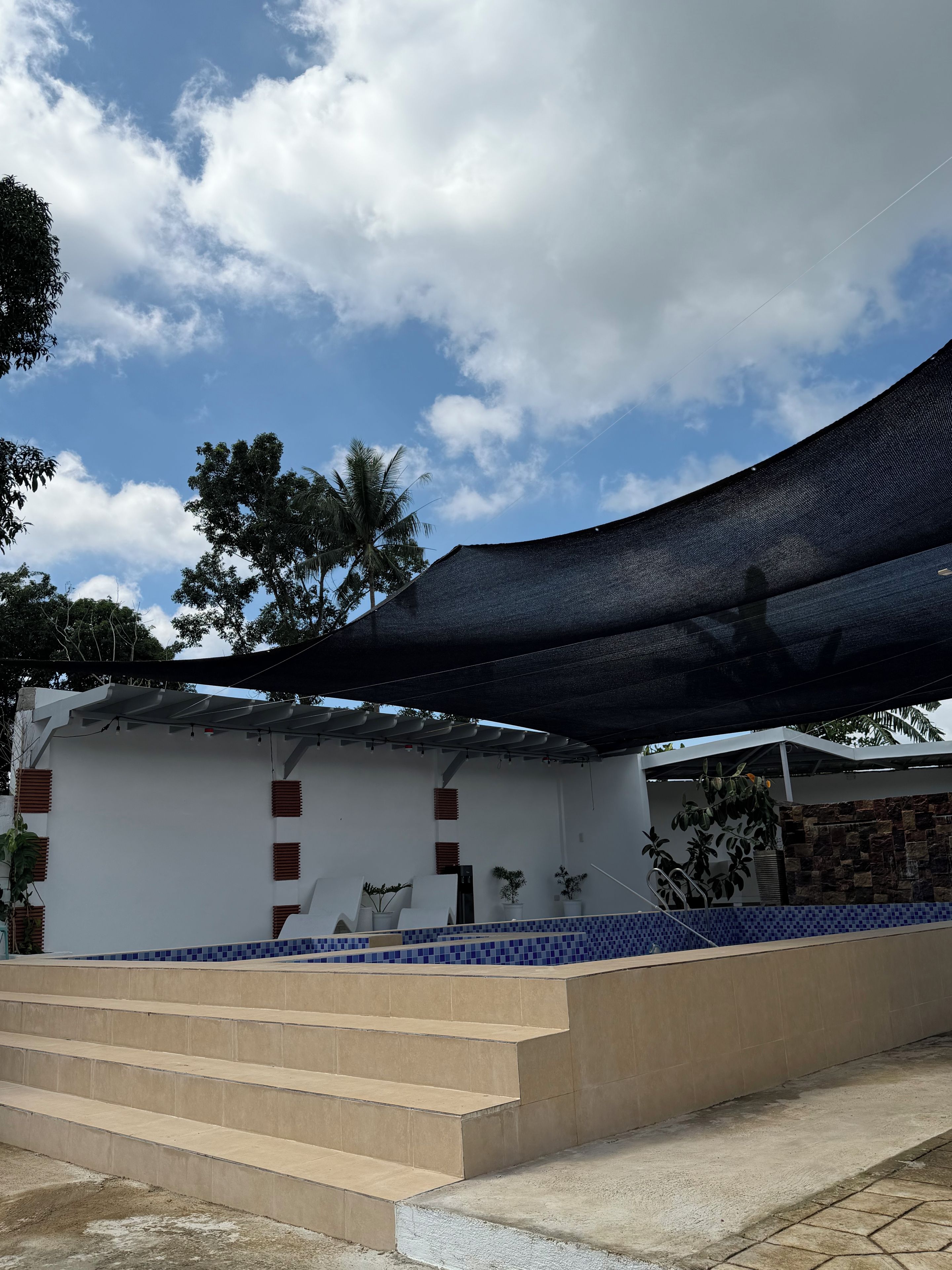 Un edificio de fachada blanca, un área de piscina con azulejos azules y una serie de escalones color beige que conducen a la piscina.