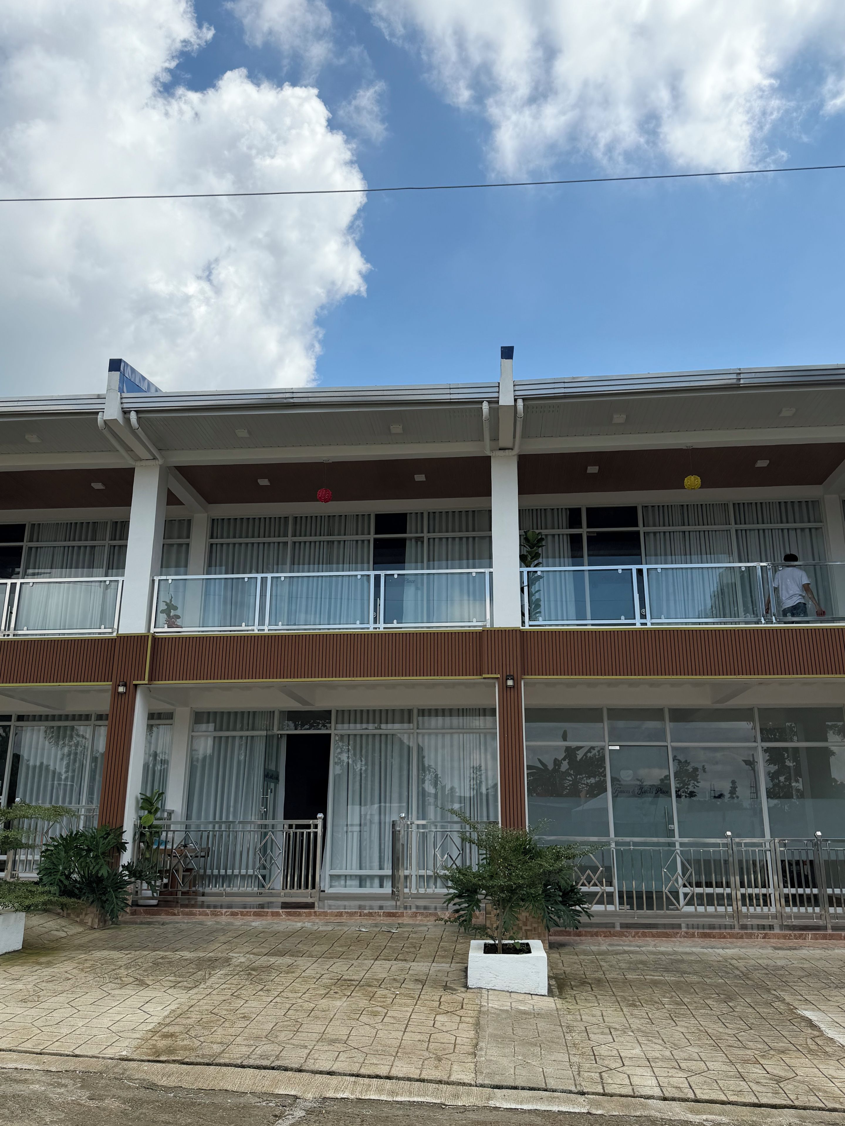 A two-story building with large windows, a balcony, and a paved entrance area.