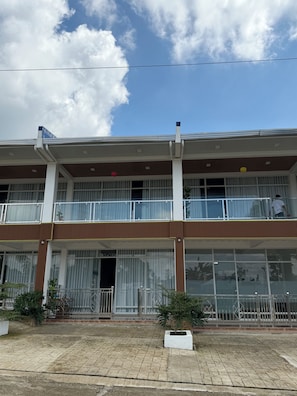 Un edificio de dos plantas con ventanas amplias, un balcón y un área de entrada pavimentada.