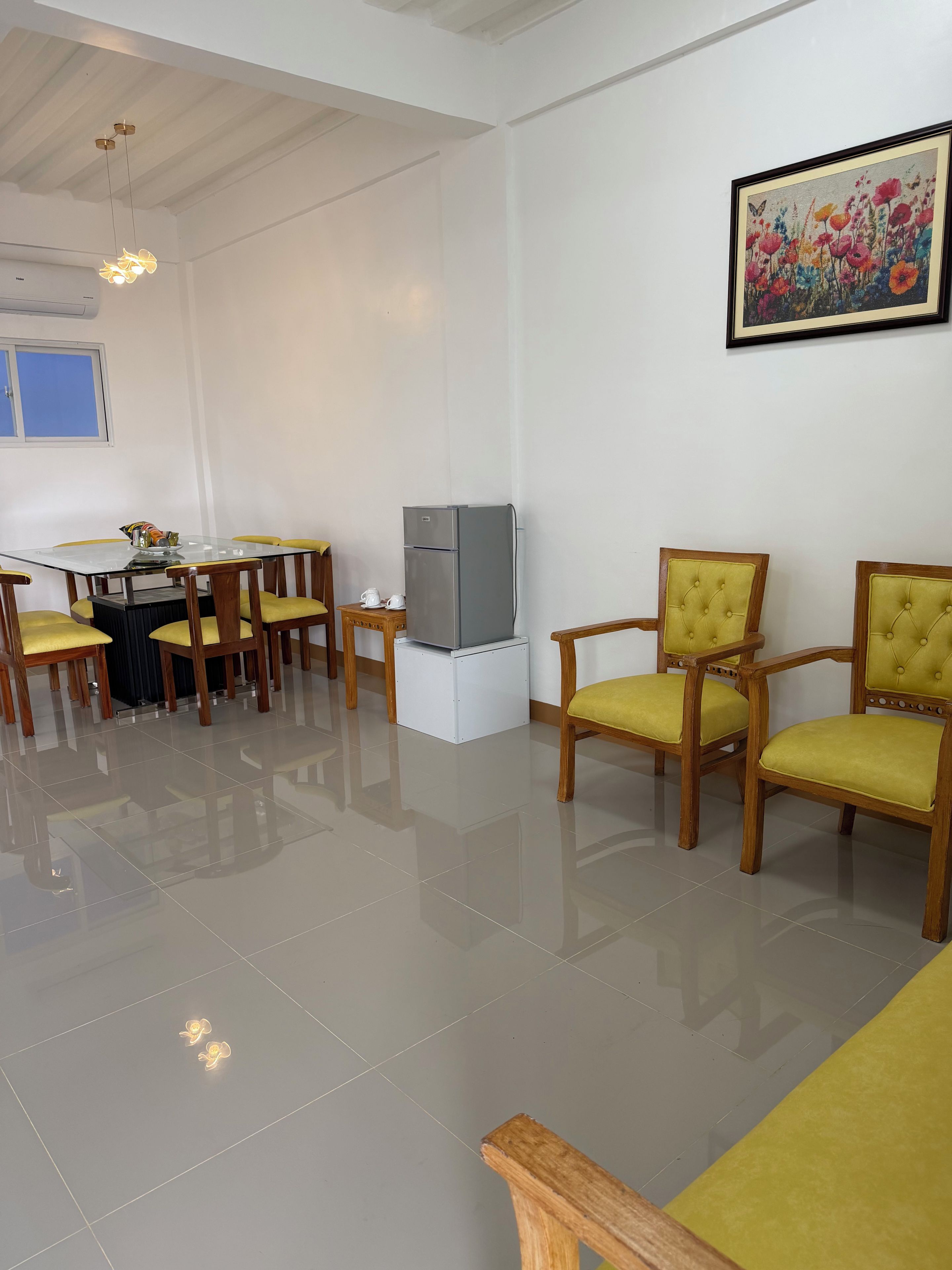 A dining area with a glass table, yellow chairs, a refrigerator, and a painting on the wall.