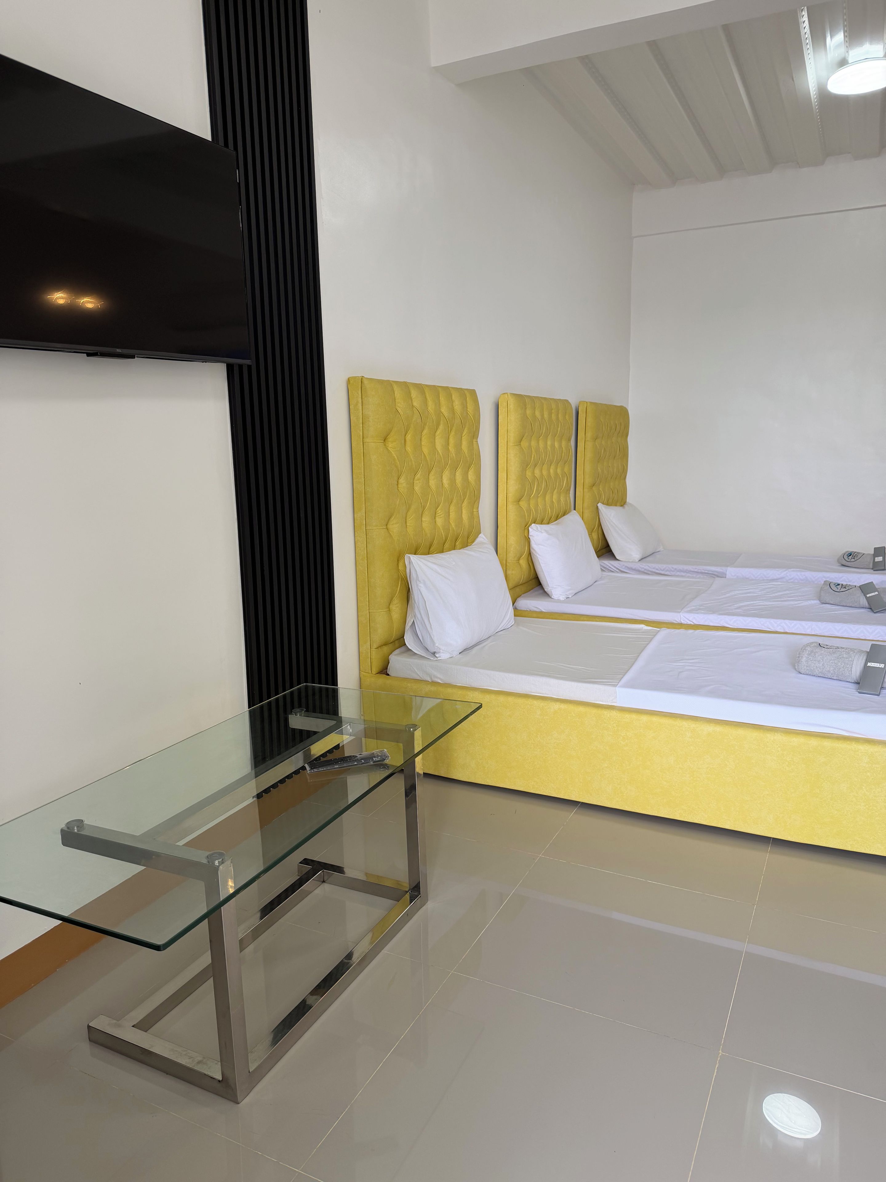 A modern hotel room with a glass table, two beds with yellow headboards, and a white wall.
