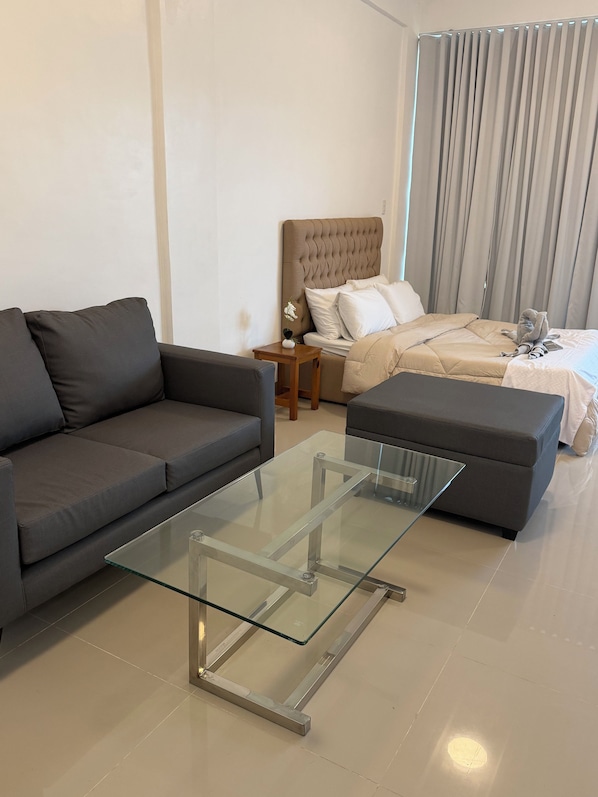 Un salon moderne avec une table basse en verre, un canapé, un lit avec une tête de lit et une table de chevet avec une lampe.
