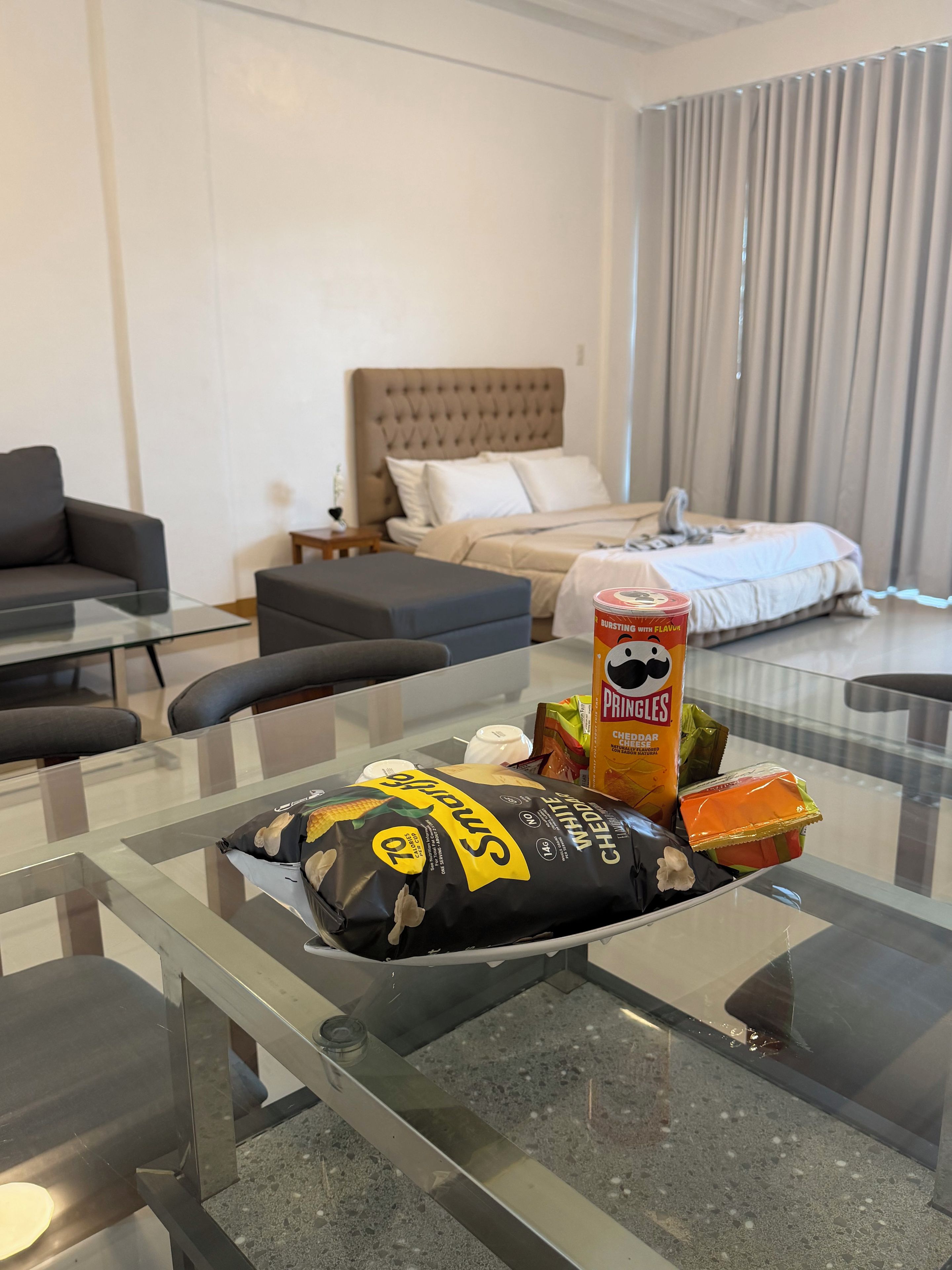 A hotel room with a bed, a sofa, a glass coffee table with snacks, and a view of the outside through the window.