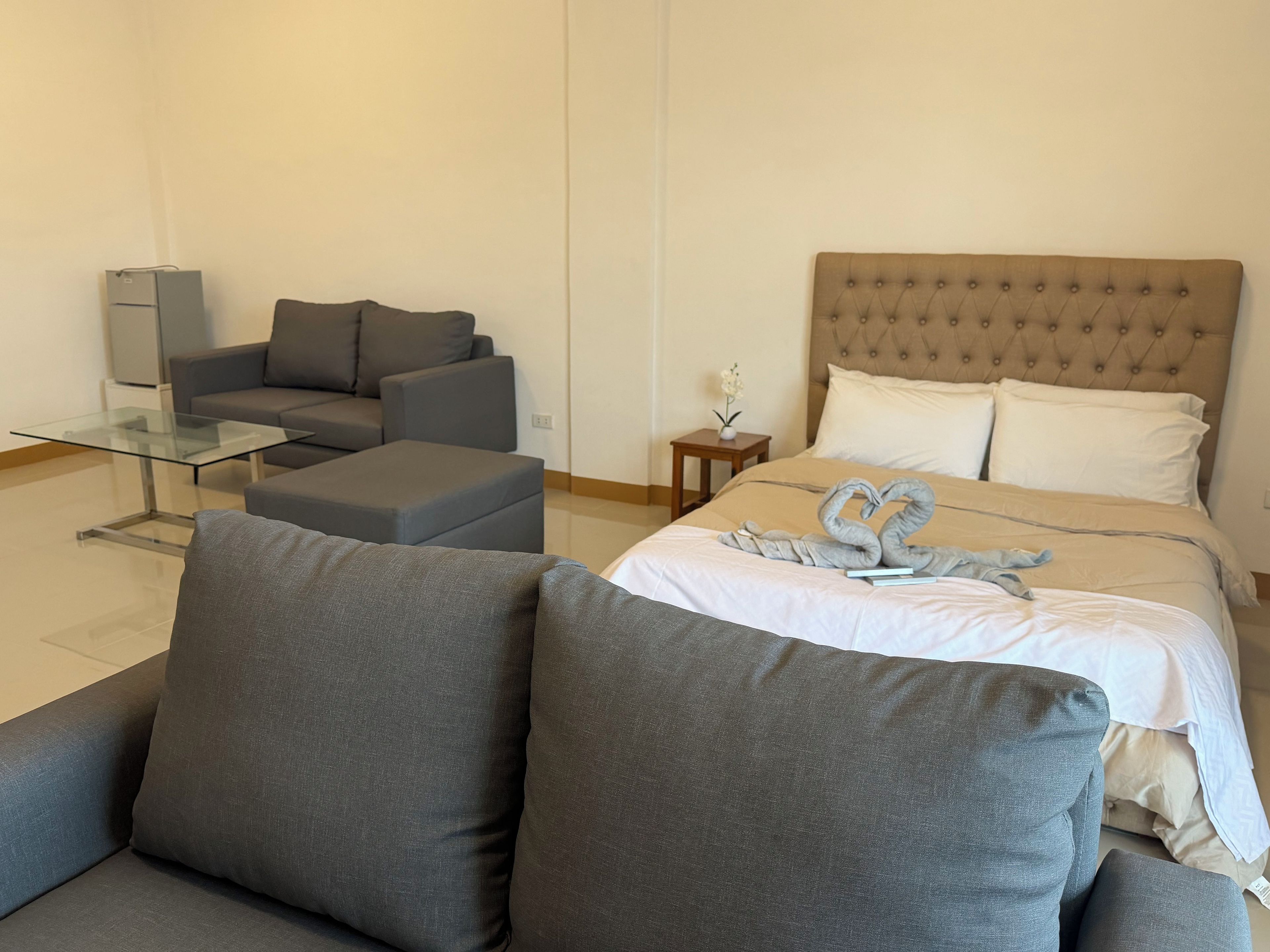 A modern hotel room with a sofa, a glass coffee table, a bed with a tufted headboard, and a decorative swan towel arrangement.