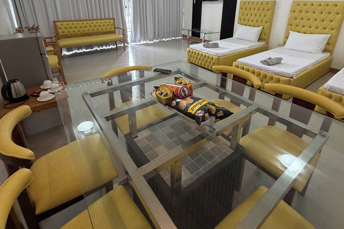 Habitación de hotel moderna con una mesa de comedor de vidrio, sillas tapizadas en color amarillo, un sofá y dos camas con cabeceros amarillos.
