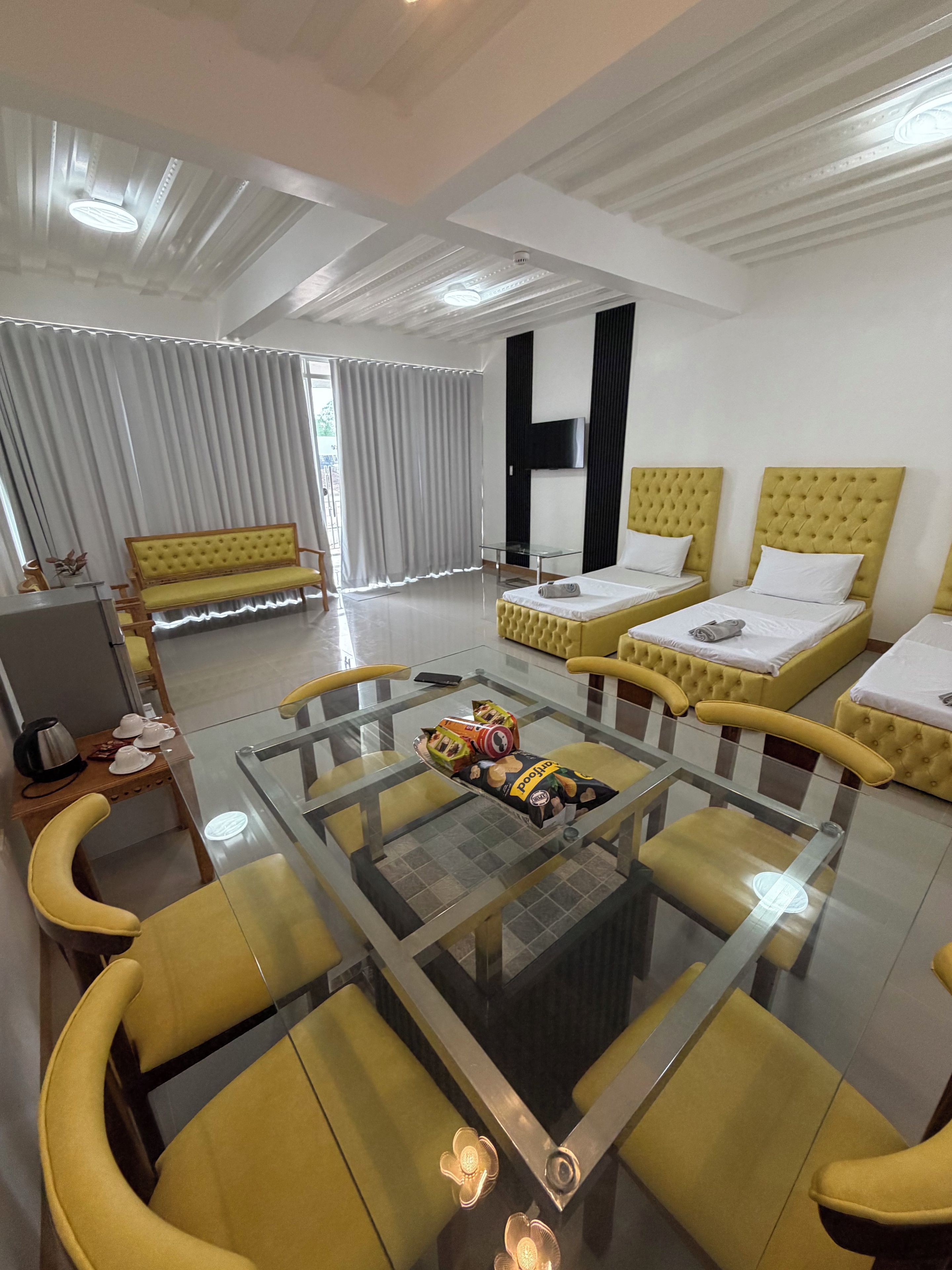 Habitación de hotel con una mesa de centro de vidrio, sillones tapizados de color amarillo, un sofá, dos camas con cabeceros amarillos y un televisor montado en la pared.