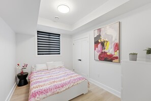 Ein Schlafzimmer mit Bett, einem Fenster mit Jalousien, einer Tür, einem Wandbild und einer kleinen Topfpflanze.