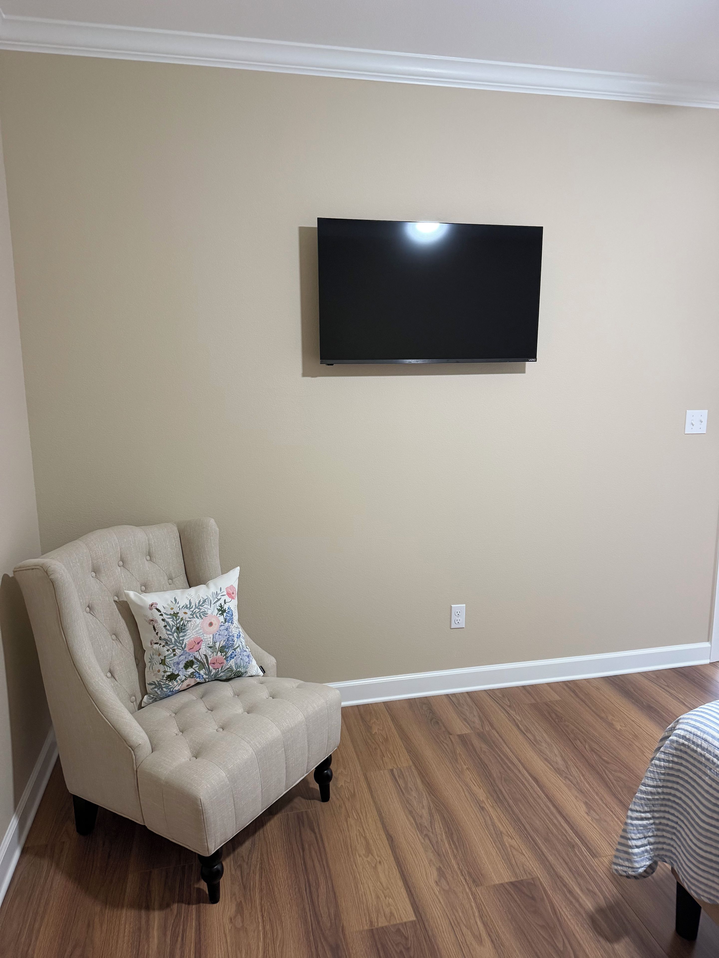 A living room with a beige wall, a flat-screen TV mounted above a beige armchair with a floral pillow, and a wooden floor.